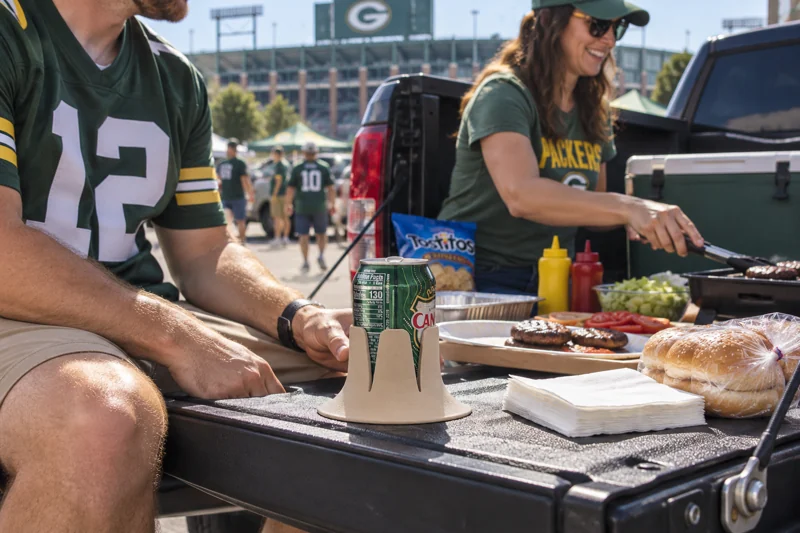 Adults preparing stadium parking-lot food at a parked pickup with opened 12-ounce soda can in Steadi on level lowered pickup tailgate with full base support next to seated adult. AI illustration.